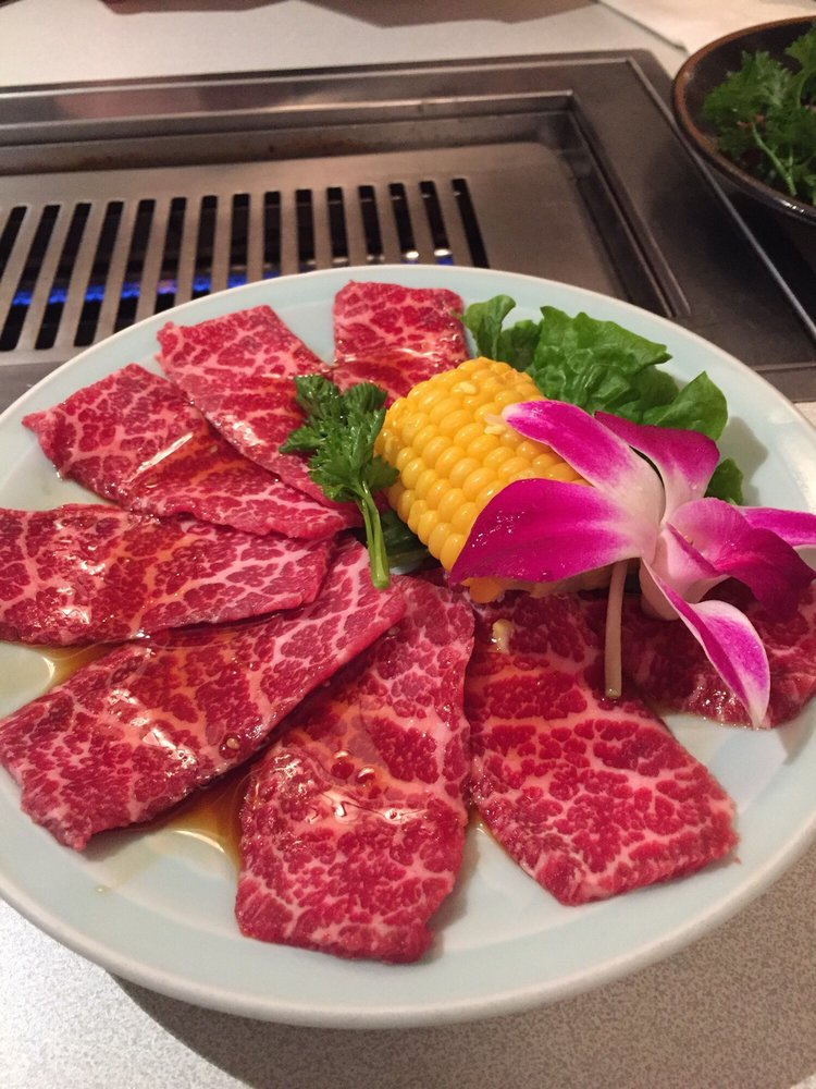 Marbled prime beef slices on a plate with orchid garnish, charcoal grill visible in background